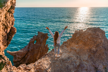 Fototapeta premium A female tourist, standing with her hands up, reaches the ends of the earth and sees the dragon rock. The girl welcomes the sun. Concept design. A successful woman looks at the sunset with open arms.