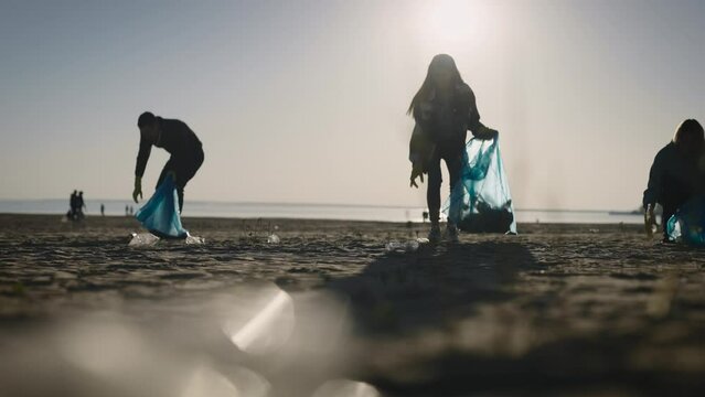 A group of eco-activists collects and sorts garbage on the beach. Silhouettes of a man and a woman against the backdrop of the setting sun.