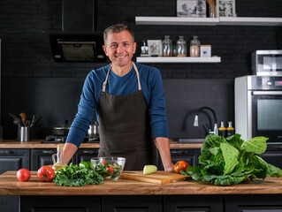 Smiling  man cooking salad, cutting fresh vegetables in modern black kitchen, happy satisfied male preparing vegetarian  food for dinner. Cabbage, tomatoes, zucchini, pepper on the back.