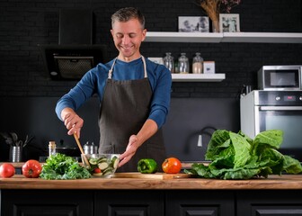 Smiling  man cooking salad, cutting fresh vegetables in modern black kitchen, happy satisfied male preparing vegetarian  food for dinner. Cabbage, tomatoes, zucchini, pepper on the back.
