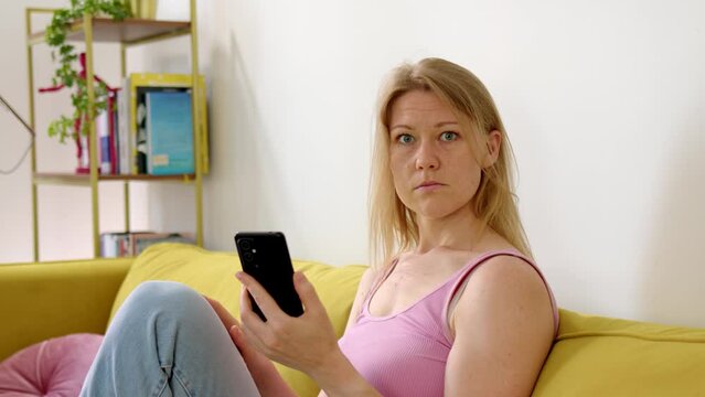 Surprised and frustrated woman looks into phone and shows it to camera. Chromakey green screen on phone to insert mockup, video or image. Incomprehensible emotions of blonde sitting at home on couch.