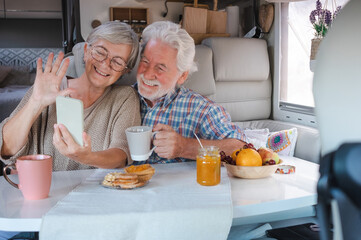 Happy senior couple sitting inside a camper van in video call with mobile phone, elderly retirees in alternative lifestyle enjoying breakfast together