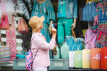 Elderly woman in hat holding credit card while looking at clothing store window with offers, ready to go in to buy and spend - consumerism concept