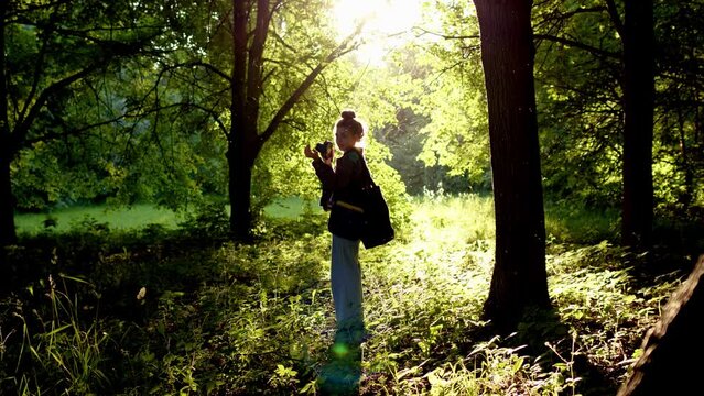 professional photographer chooses place for photo session in rays of setting sun, which creates excellent backlight on person. Woman in with camera stands in forest against backdrop of dawn sun