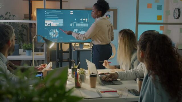 Office worker pointing at digital screen and discussing marketing ideas in boardroom at night. Young lady making presentation for creative team.