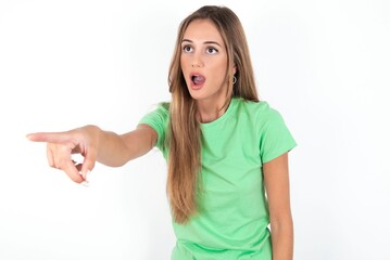 young beautiful woman wearing green T-shirt over white background Pointing with finger surprised ahead, open mouth amazed expression, something on the front.