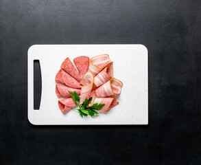Food meat assortment bacon sausage salami slices on a cutting white board on a black background,