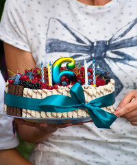 A festive cake decorated with different types of berries, cookies and a multi-colored number six in the hands of the mother
