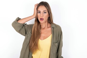 Embarrassed young beautiful woman wearing green overshirt over white background with shocked expression, expresses great amazement, Puzzled model poses indoor