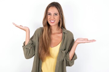 Indignant young beautiful woman wearing green overshirt over white background gestures in bewilderment, frowns face with dissatisfaction.