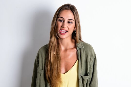 Funny young beautiful woman wearing green overshirt over white background makes grimace and crosses eyes plays fool has fun alone sticks out tongue.