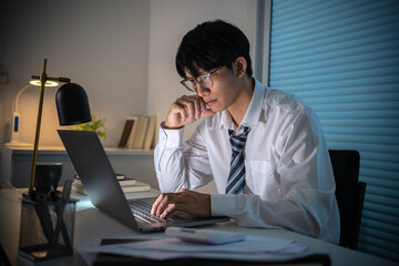 Alone Working Late at Night.Asian business man at workplace in office holding his head on hands after late night work at a desk in a dark office working on a laptop late in the evening concept