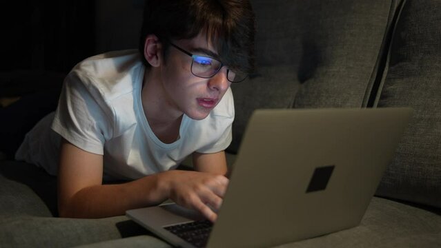 Teenager nerd boy typing on laptop. Tired cute boy working at night laying on couch. Overworking concept
