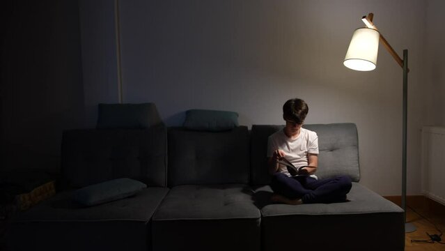 Teenager boy reading or taking a look at book at night sitting on couch in the living room