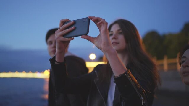 Friends are sitting on the shore, a young woman takes pictures on the phone. Embankment, summer night. Slow motion 4k footage