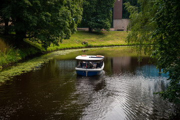 A tourist boat takes tourists on a river tour in the canal city. River with water lilies and green trees on its bank.