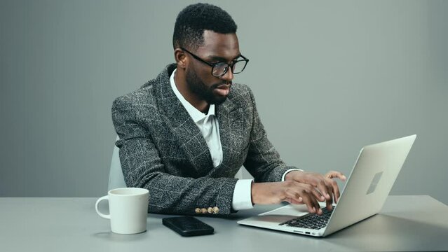 African American businessman working in the office at a laptop with a mug of coffee at his desk on a gray background and tired