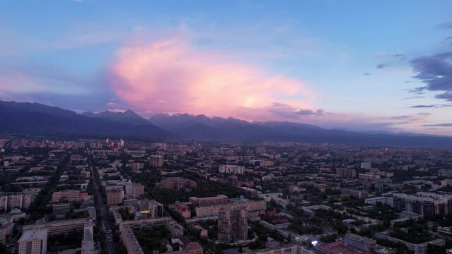 Pink and white sunset, clouds over the mountains and the city of Almaty. Huge fluffy clouds along the mountains. The houses were painted pink. Cars are driving on the roads. There is Kok-Tobe TV tower