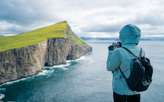 Back view of woman wearing winter clothes taking pictures Sorvagsvatn lake on Vágar Island. Tourist popular attraction destination in Faroe Islands. Young tourist woman taking photo with smart phone. 