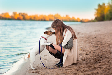 A girl walks with a dog on the river bank. The dog licks the girl's face. Autumn trees are visible in the distance.