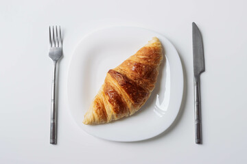 a freshly baked croissant lies on a white plate with a fork and a knife on the sides close-up isolated shot on a white...