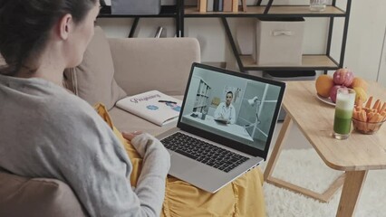 Pregnant woman speaking with female doctor via video call on laptop during online consultation while sitting on sofa at home by table with healthy snacks and green smoothie on it