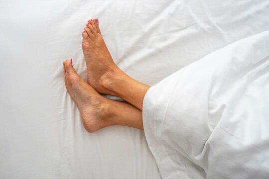 Woman legs on the bed sheet under blanket top view close-up. Female bare feet on a white sheet, top view, closeup. Closeup naked female legs model onthe bed
