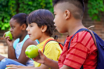 multiethnic kids with backpacks sitting on the street at school entrance eating apples and talking...