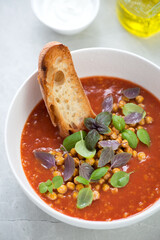 Close-up of tomato and chickpea soup served with fresh basil and ciabatta in a white bowl, vertical shot on a light-grey stone surface