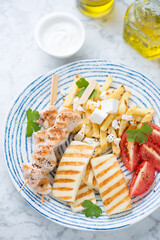 Plate with slices of grilled haloumi cheese, souvlaki skewers and potato fries, vertical shot on a light-grey marble surface
