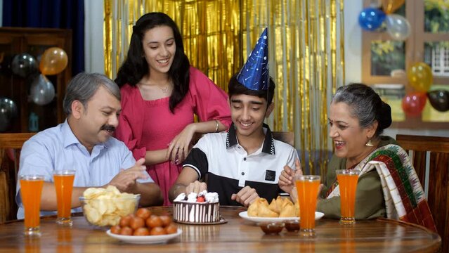 A handsome teenage boy wearing a birthday cap blowing birthday candles and cutting a birthday cake - home celebration. Happy Indian family - Small nuclear family celebrating at home  party time  fu...