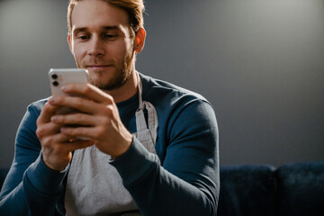 Young man wearing apron using cellphone while sitting on sofa at home