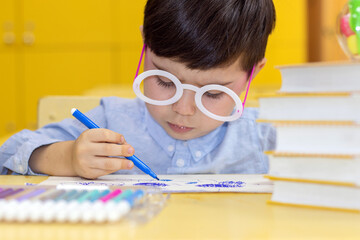 Naklejka na ściany i meble preschool kid sits in school desk reading looking in book or drawing with colorful markers.serious face child wearing party round glasses frame with no lens.smart little boy first grade back to school