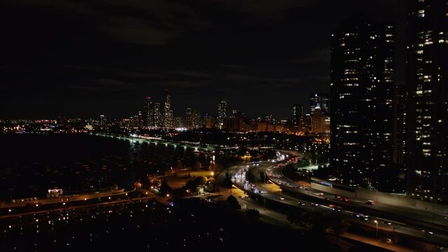 Aerial drone shot of traffic movement over a bridge along the seaside with the view of high-rise office and residential buildings in the background in Chicago, USA at night time.