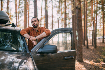 A young man stands next to a car with an upper tourist trunk in a pine forest on the shore of a lake at sunset.