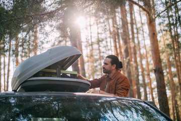 A young man opens the upper tourist trunk of a car in a pine forest on the shore of a lake at...