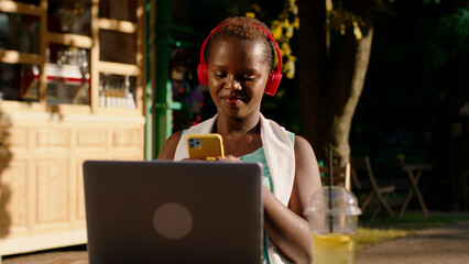 A very astonishing woman is sitting at a table in the park, smiling as she is typing something on the laptop