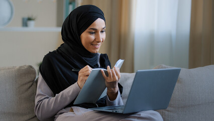 Cheerful young muslim arabian lady young woman sit on cozy sofa female freelancer writer take notes in diary noting information write book daily tasks secrets ideas in personal organizer using laptop