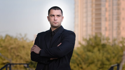 Portrait of confident strong caucasian businessman adult brunet man guy wears formal black suit posing with crossed arms outside handsome male professional boss downtown office skyscraper background