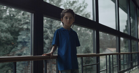 Upset schoolboy posing alone near panorama window. Lonely boy standing corridor 