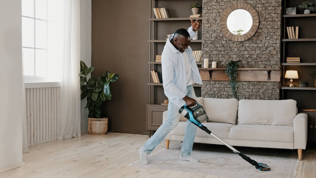 Adult african american homeowner cleaner washing floor parquet surface with vacuum cleaner in dwelling living room cleaning dust. Active elder bachelor moves to music at home doing housekeeping chores
