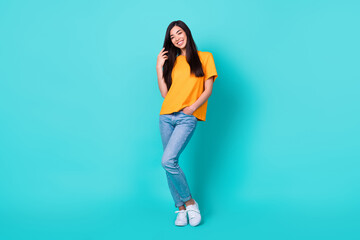 Fototapeta na wymiar Full body photo of sweet brunette lady touch hair wear orange t-shirt jeans sneakers isolated on turquoise color background