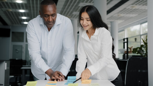 Two colleagues african and arab businesspeople co-workers writing tasks on sticky notes brainstorming organizing work together managing paperwork in office using colorful memo stickers show thumb up