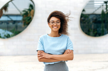 Portrait of a fashionable elegant beautiful African American young woman with curly hair, with glasses, wearing a blue shirt, standing outdoors with her arms crossed, looking at the camera, smiling