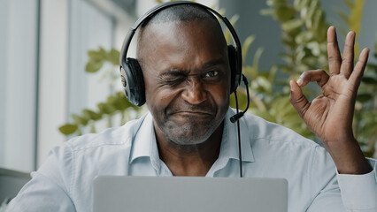 Concentrated african american senior entrepreneur businessman freelance worker wears headphones with microphone chatting online browsing internet webinar using computer app show okay recommend gesture - Powered by Adobe