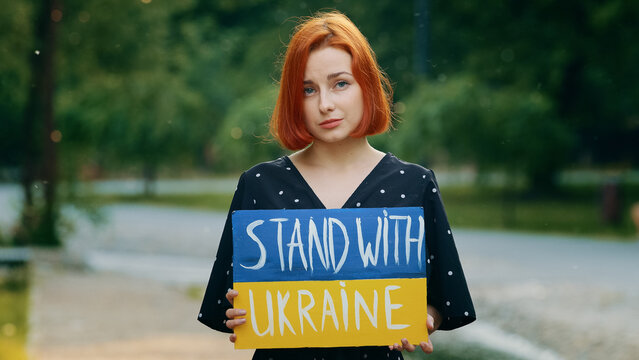 Sad redhead girl hold cardboard with written inscription phrase stand with Ukraine shows patriotic banner painted in ukrainian national blue yellow colors upset anxious young woman ask active support