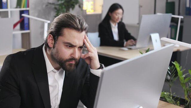 Concentrated caucasian male in formal wear sitting at office workplace and touching forehead with hand. Handsome man getting lost in thoughts during work.