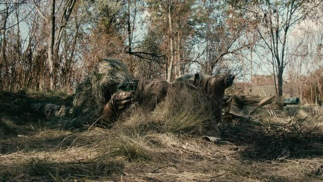 A close-up of a sniper wearing a camouflage suit or grass cloak, waiting and looking through a rifle scope. The sniper aims and shoots. Marksmanship shooting. Cinematic shot