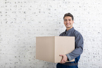 Confident smiling male from moving service holding box beside white brick wall background 