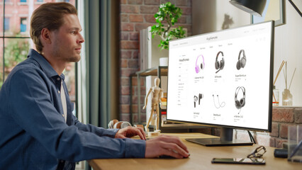 Fototapeta na wymiar At Home Office: Man Using Desktop Computer, Online Shopping for Electronics, Wireless Hi-Fi Headphones. e-Commerce Concept of Purchasing, Buying, Ordering Tech Devices on Website. Over Shoulder View